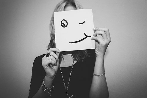 Frau versteckt sich hinter einem Blatt Papier mit  zwinkerndem Smiley