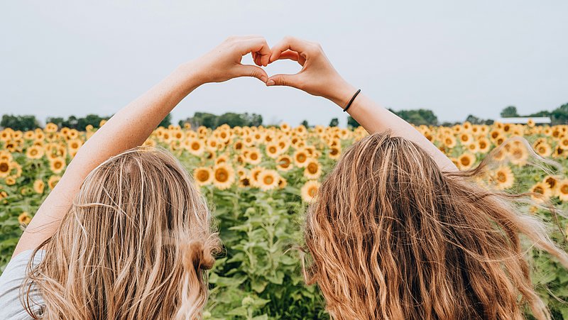 2 Freundinnen sind von hinen auf einer Blumenwiese zu sehen. Sie formen ein Herz aus ihren Händen